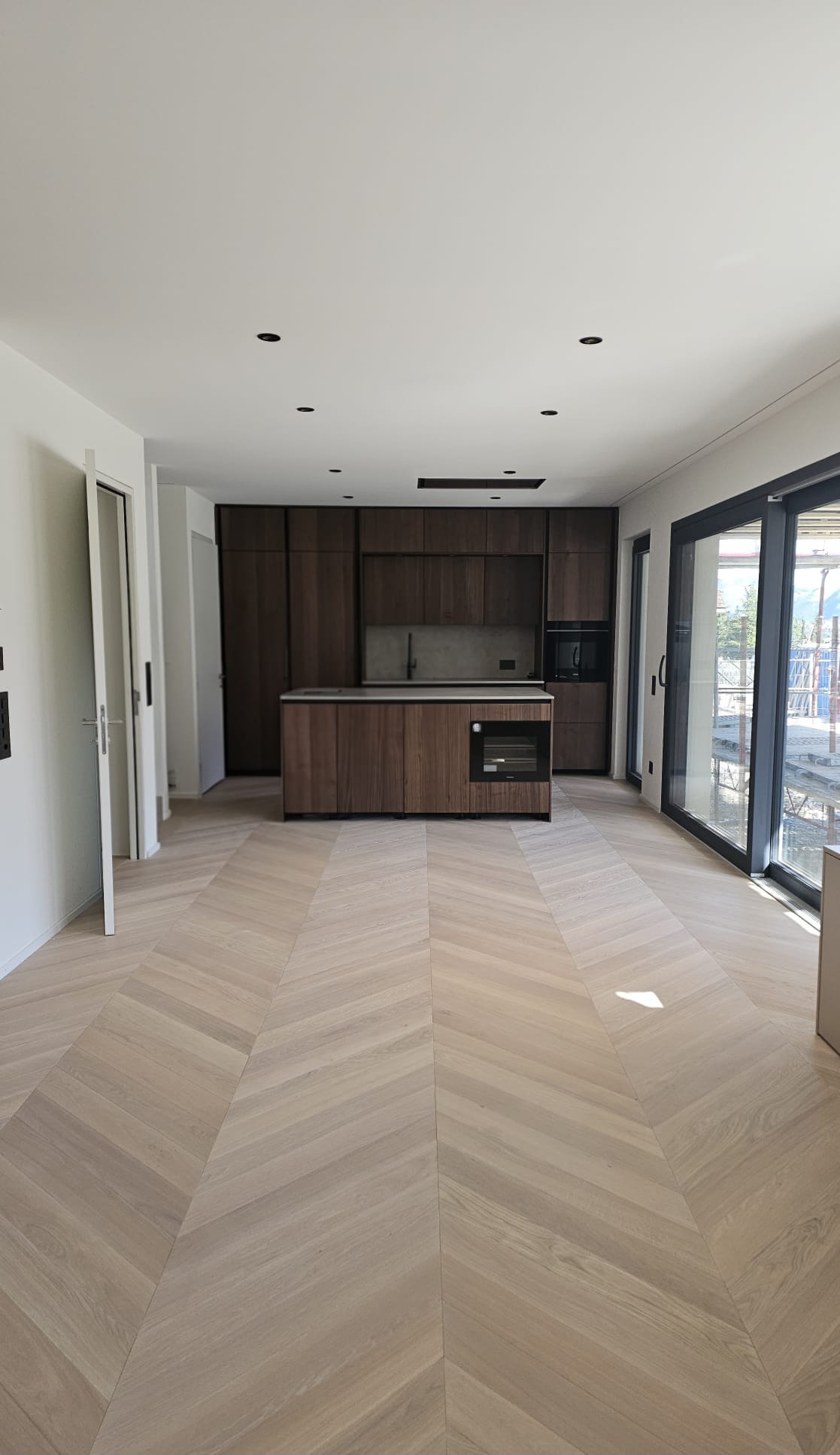 Parquet en bois clair posé en point de Hongrie dans une pièce de vie de nouvelle construction, cuisine en bois foncé au fond et grandes baies vitrées à droite