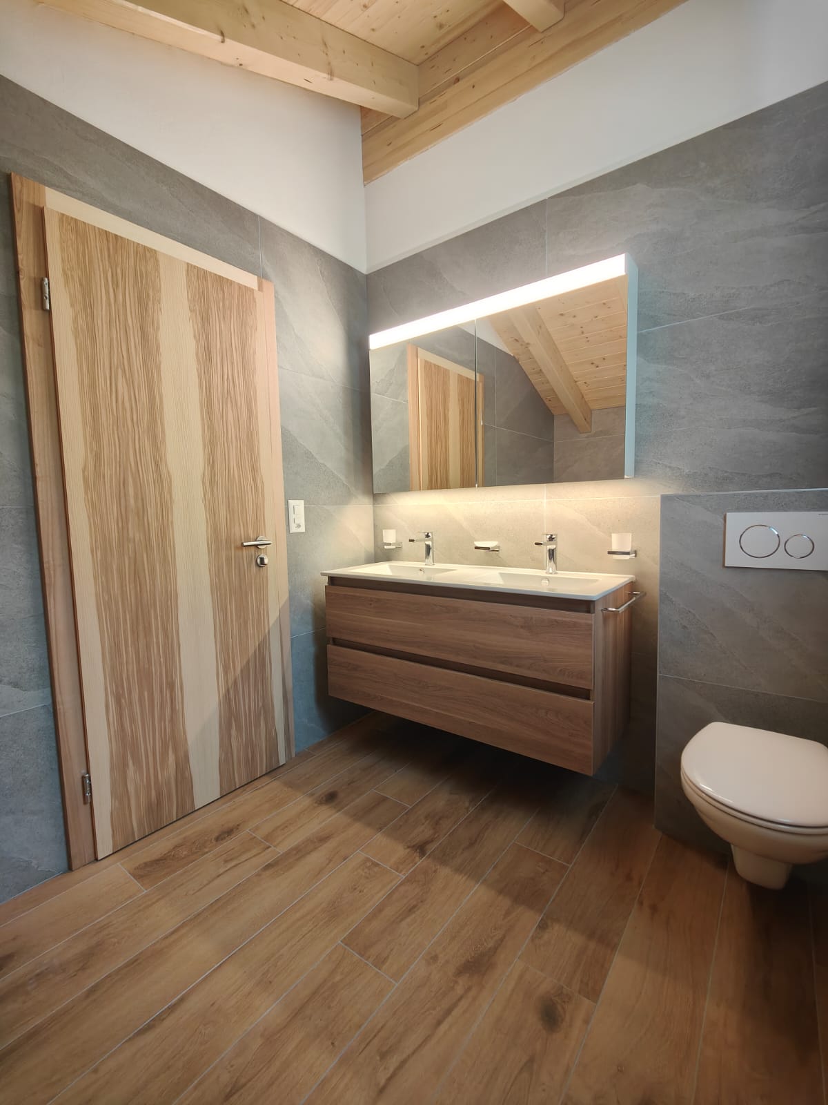 Salle de bain rénovée avec meuble double vasque en bois, miroir lumineux et carrelage effet pierre